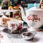 Preview: Mit Blumen verzierte Kanne und Tasse mit Teesieb im Vordergrund und rustikale Küche im Hintergrund