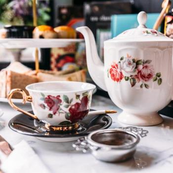 Mit Blumen verzierte Kanne und Tasse mit Teesieb im Vordergrund und rustikale Küche im Hintergrund
