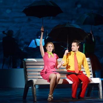 Ein Theaterstück 2 Schauspieler die auf einer Bank sitzen mit einem Regenschirm in der Hand eine Musikband im Hintergrund