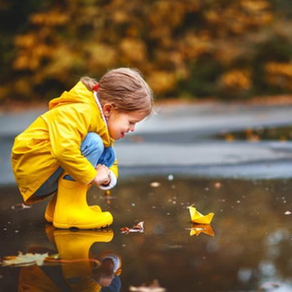 Kind was an eine Pfützemit einem Papierboot spielt in gelber Regenkleidung rustikaler Hintergrund
